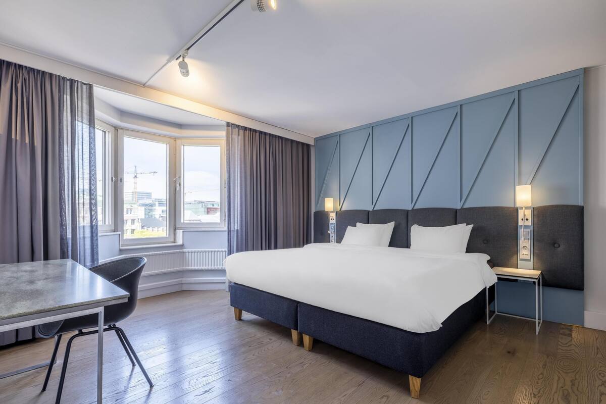 A modern hotel room with a large bed, a desk, and a view of the city through the window.