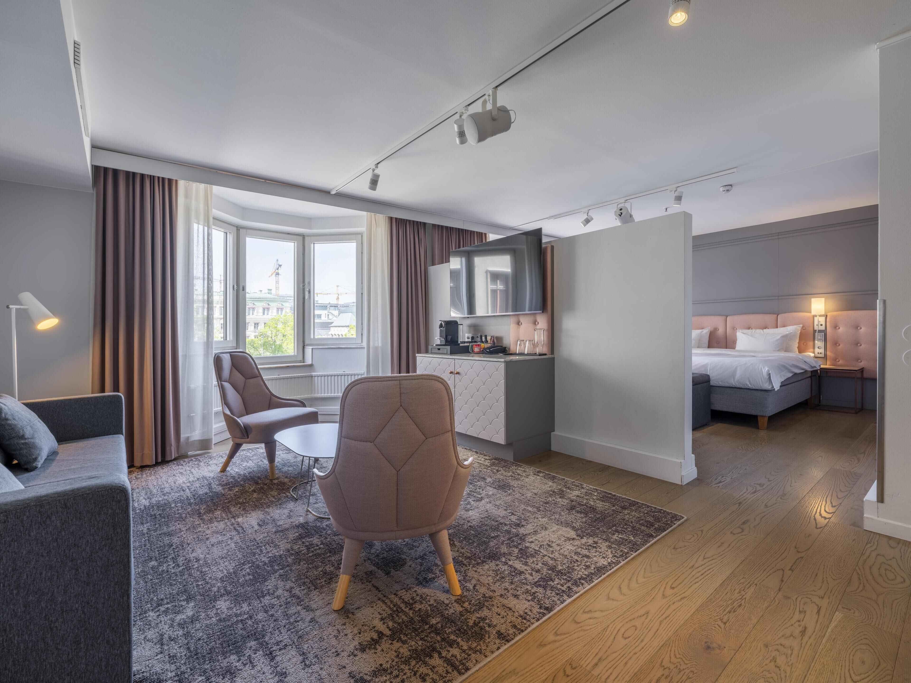 Ett modernt hotellrum med en soffa, en fåtölj, ett litet bord, en säng, ett litet kök och utsikt över staden.