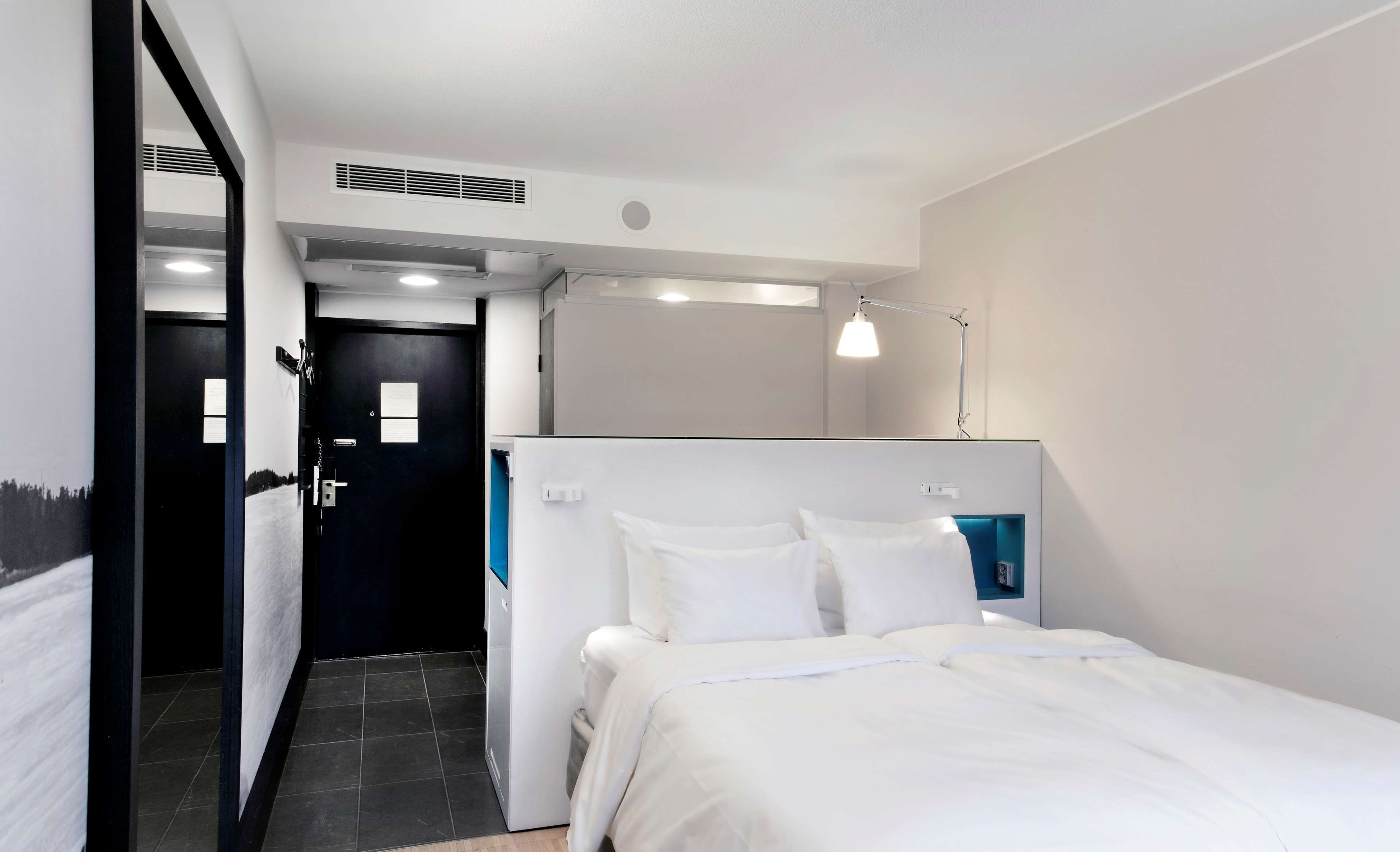 A hotel room with a bed, a desk, a chair, a desk lamp, and a door.
