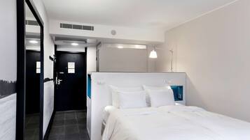 A hotel room with a bed, a desk, a chair, a desk lamp, and a door.