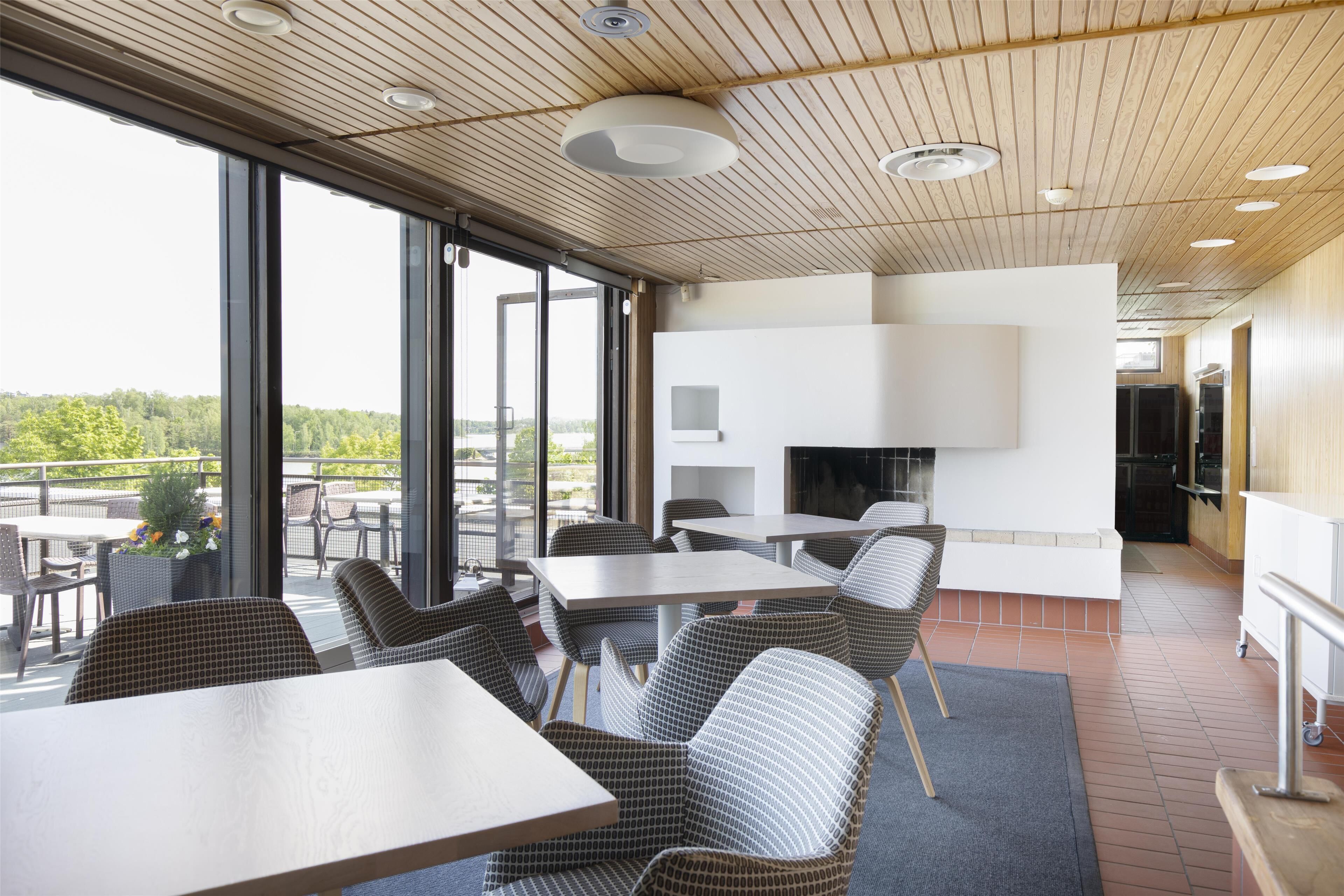 A modern dining area with large windows, wicker furniture, and a kitchen area visible through the glass.