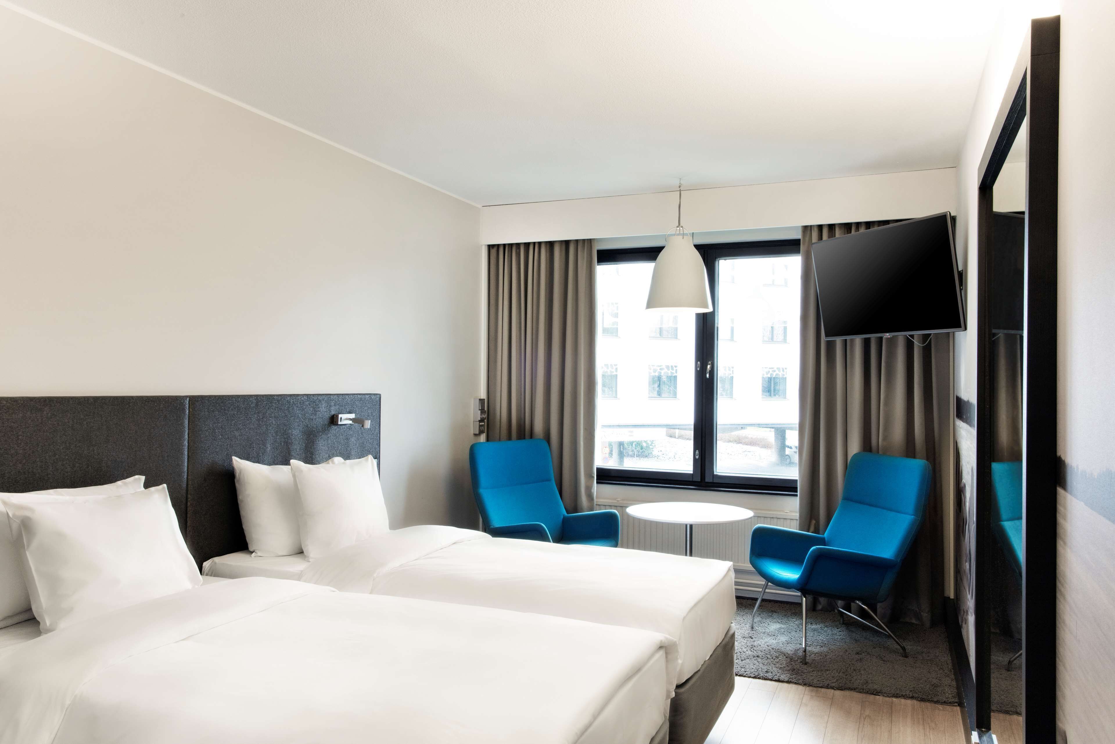 Ein Hotelzimmer mit zwei Betten, einem Fernseher, einem kleinen runden Tisch und zwei blauen Stühlen.