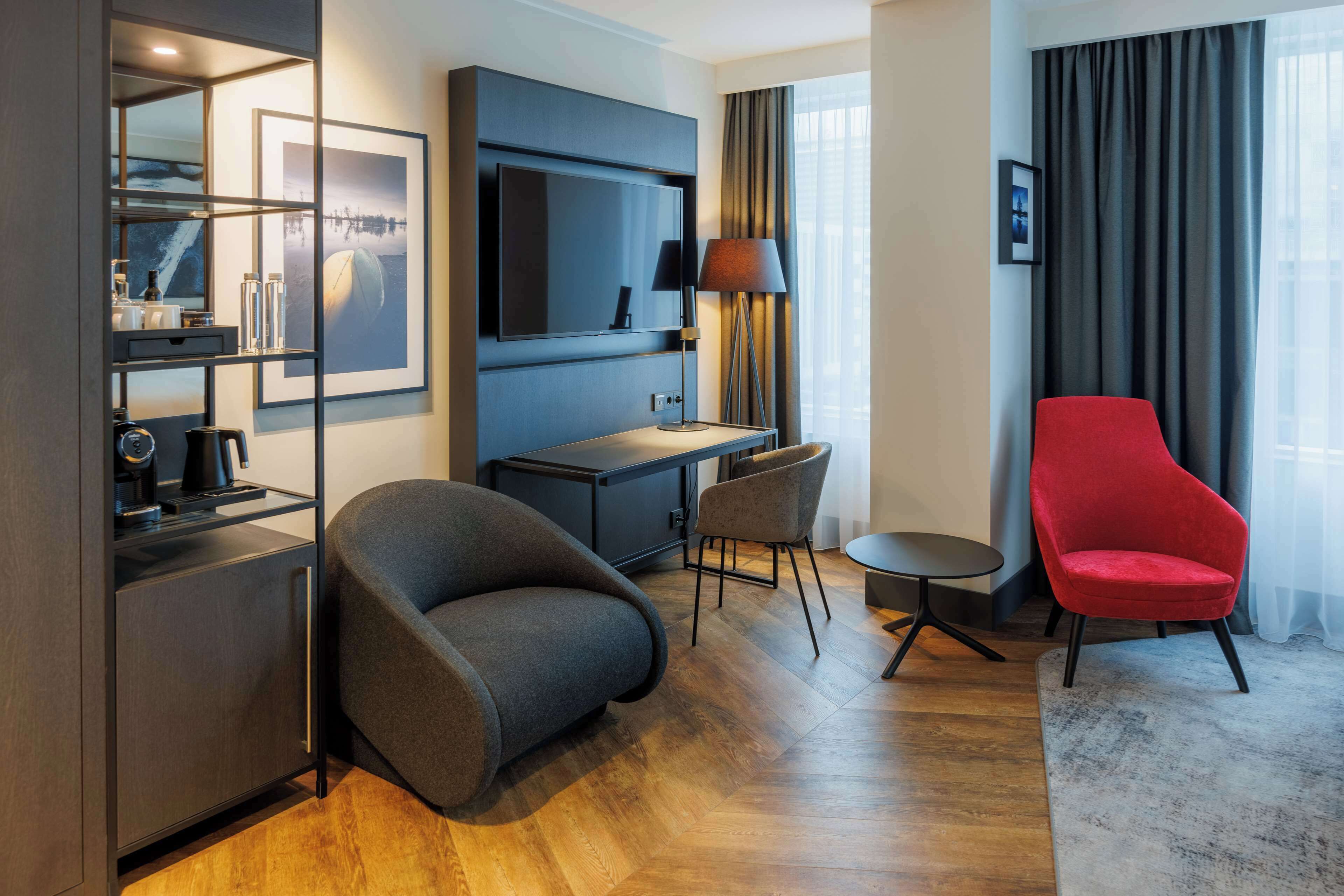 Una habitación de hotel moderna con un sofá gris, un sillón rojo, un escritorio con lámpara, televisión y un ventanal con cortinas.