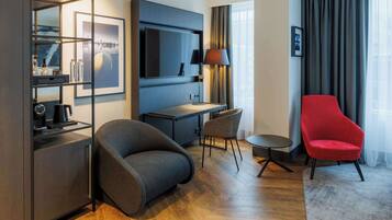 Una habitación de hotel moderna con un sofá gris, un sillón rojo, un escritorio con lámpara, televisión y un ventanal con cortinas.