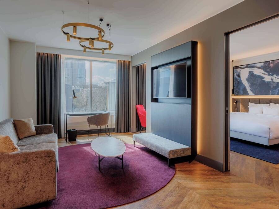 Moderni hotellihuone, jossa on sohva, pyöreä pöytä, penkki, televisio ja sänky.