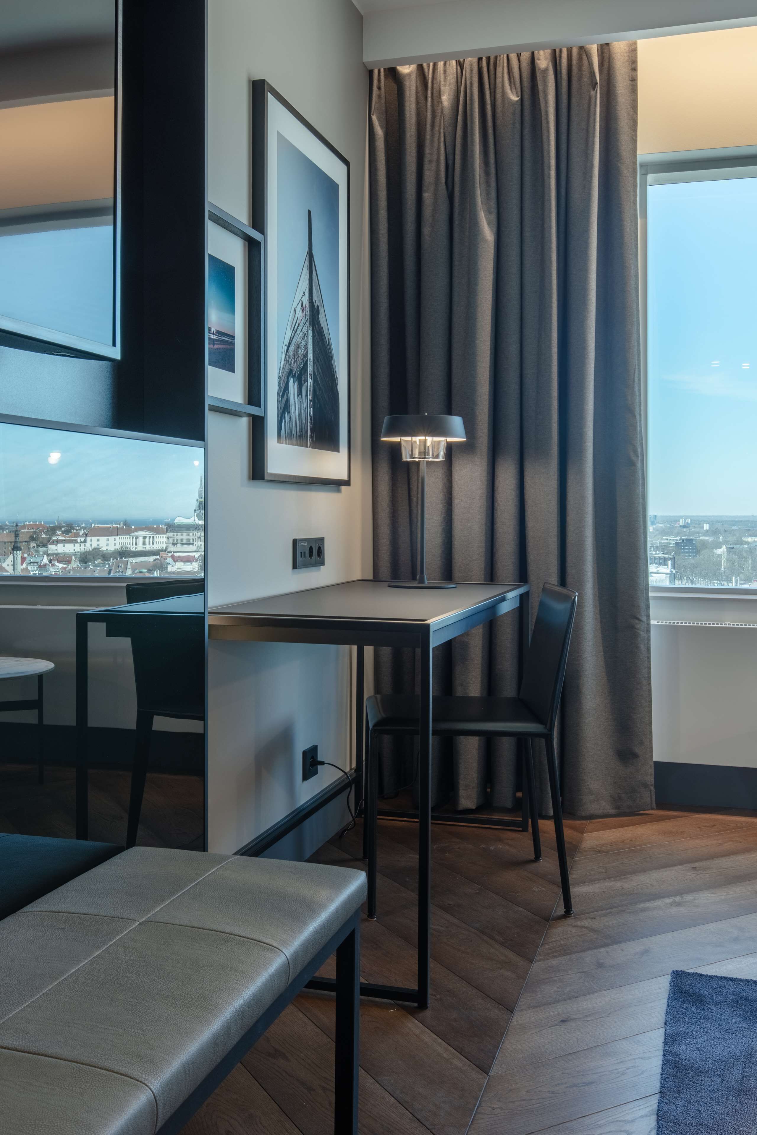 A modern hotel room with a dining area, a large window with a city view, and a desk with a lamp.