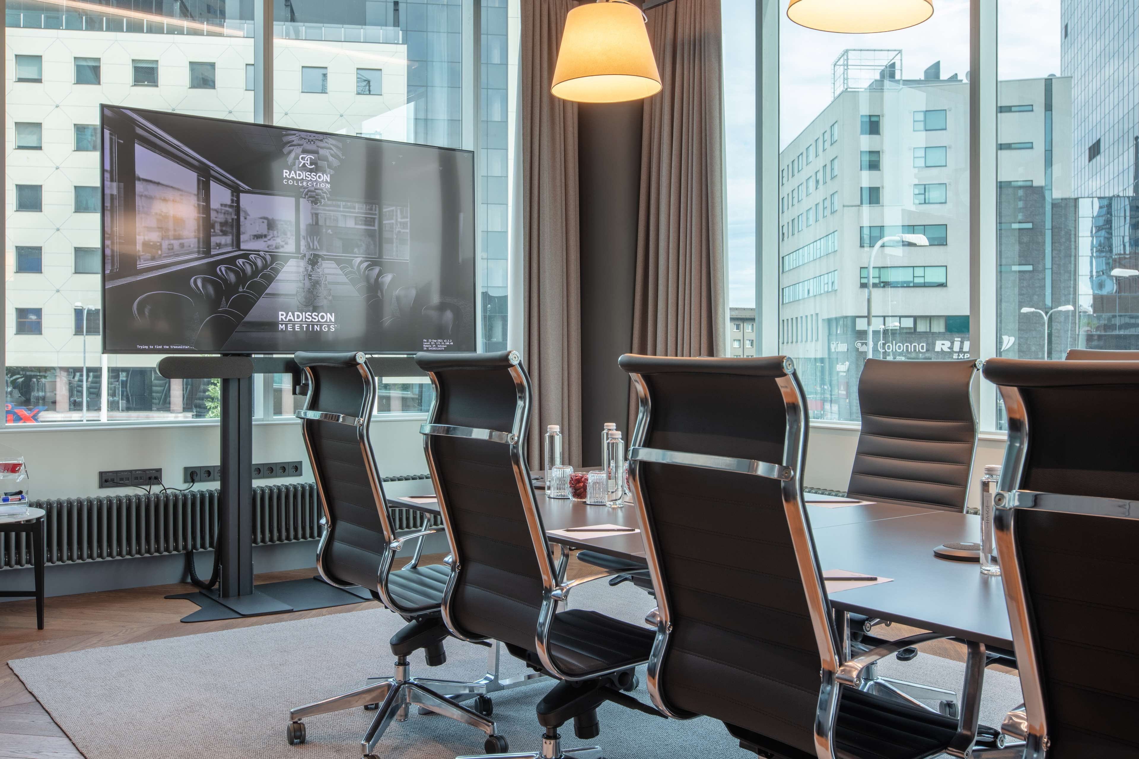 Une salle de conférence dotée d’un grand écran affichant le logo « Radisson Meetings », de quatre chaises noires à structure chromée, d’une table avec des bouteilles d’eau et offrant une vue sur les bâtiments à travers de grandes fenêtres.