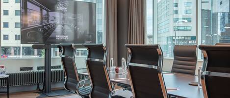 Un salón de conferencias con una pantalla grande mostrando 'Radisson Meetings', cuatro sillas negras con estructura cromada, una mesa con botellas de agua y vistas a edificios a través de amplias ventanas.