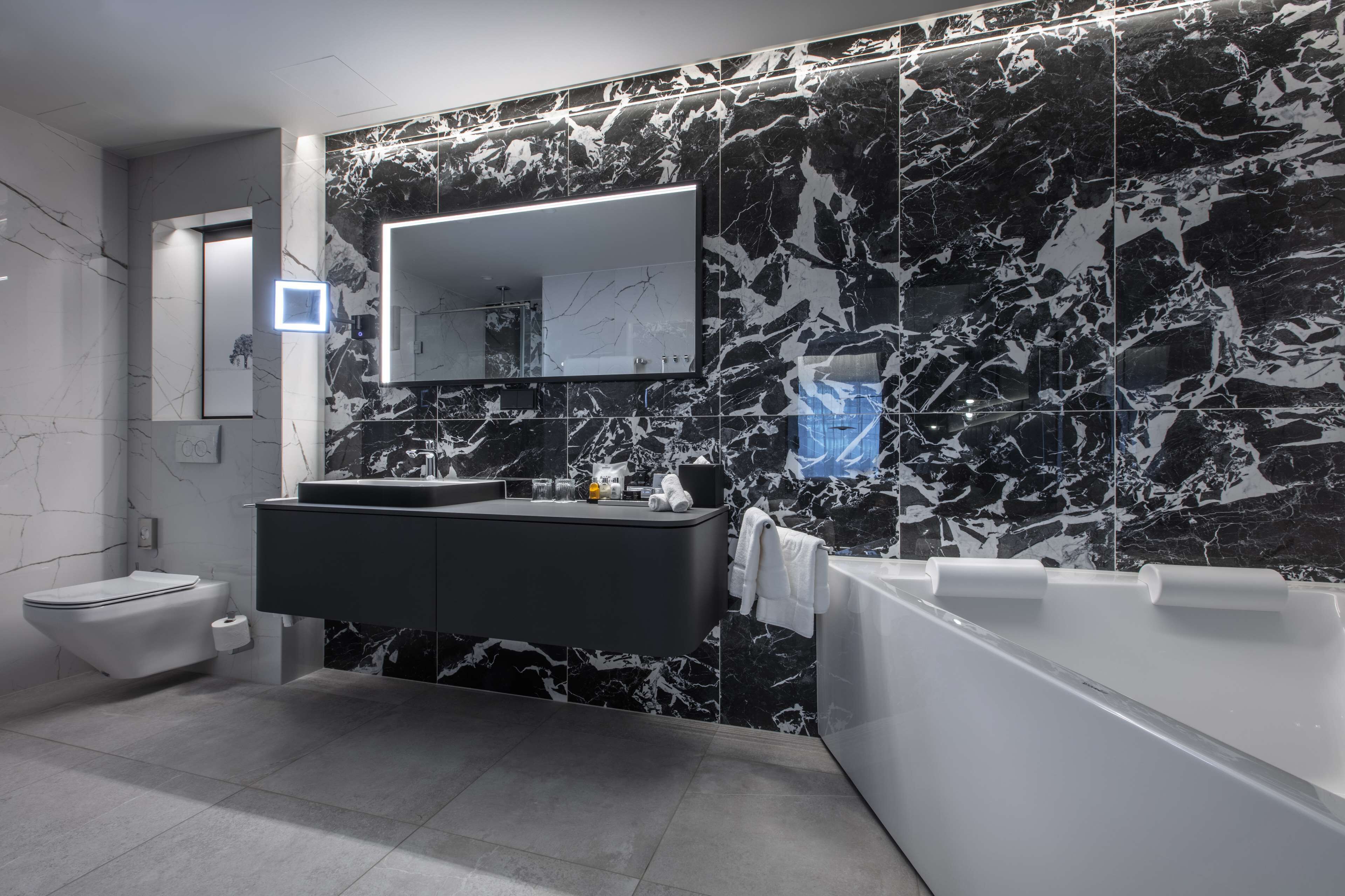 A modern bathroom with a black and white marble backsplash, a large mirror, a black vanity, a white toilet, and a bathtub.