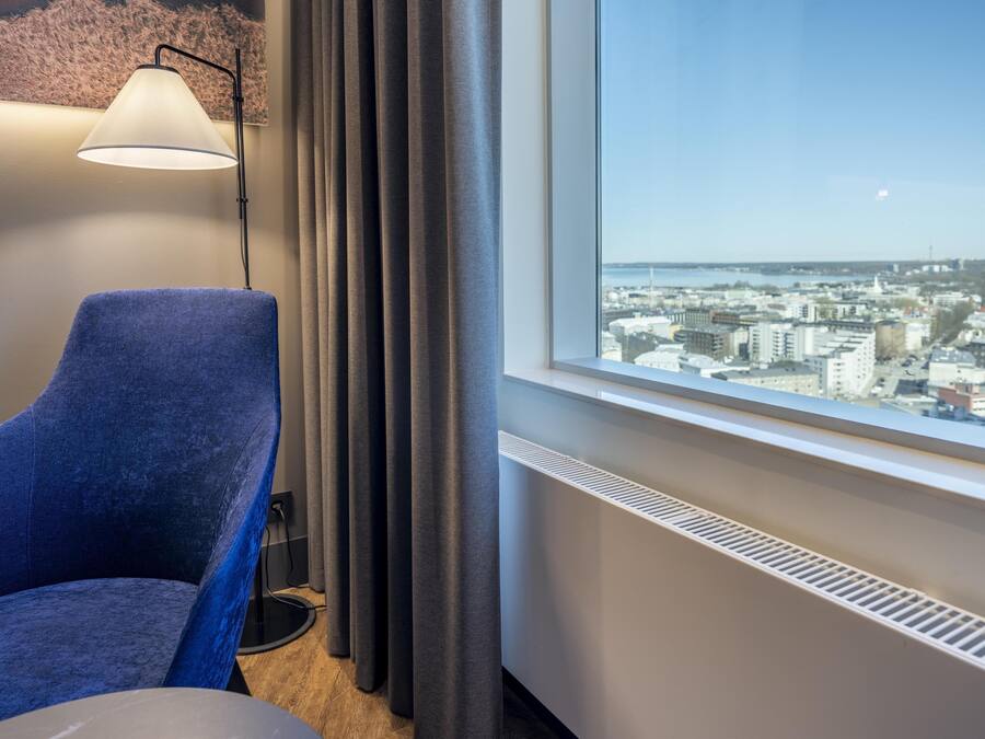 Habitación de hotel con vista a la ciudad, un sillón azul, una lámpara de pie y un radiador.