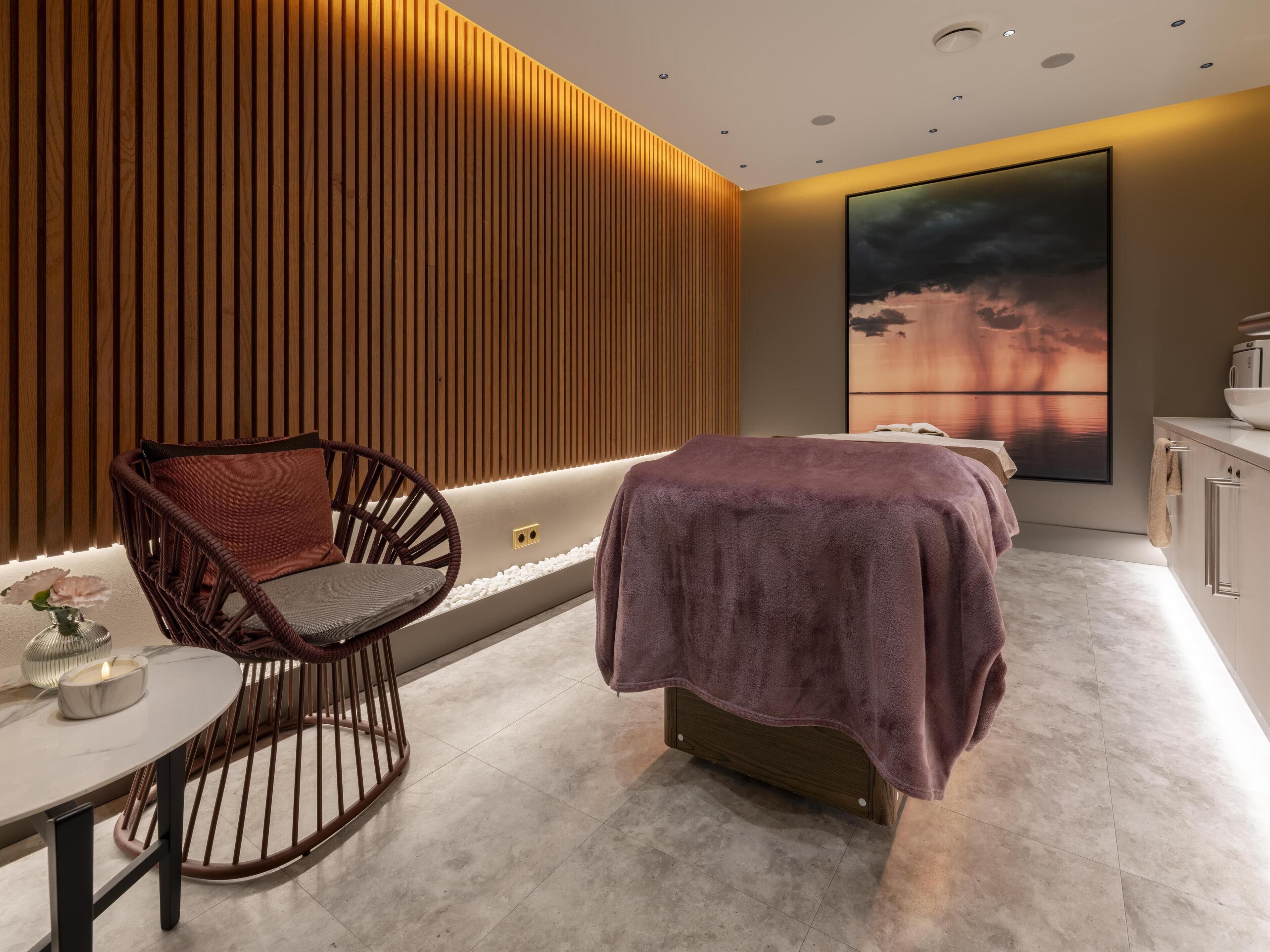 A modern spa room with a wooden slat wall, a massage table, a wicker chair, and a large artwork.