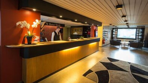 A hotel reception desk with a modern design, a receptionist, and a seating area with a large window in the background. - Q Hotel Växjö (Vaxjo)