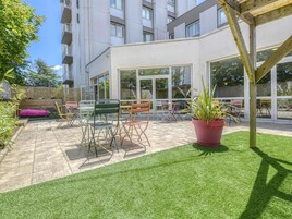 Un espace extérieur aménagé en terrasse, doté d’une pergola en bois, d’un gazon synthétique vert et de différents modèles de chaises et de tables en métal.