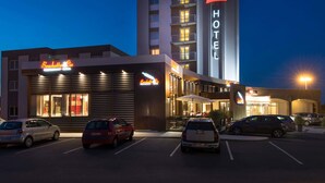 Un hotel amb un cartell destacat de 'ibis HOTEL', un aparcament amb diversos cotxes i una botiga anomenada 'Brochettes & Co'.