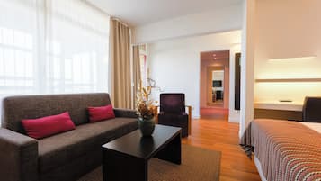 A modern hotel room with a grey sofa, a small table, a chair, and a bed with a patterned bedspread.
