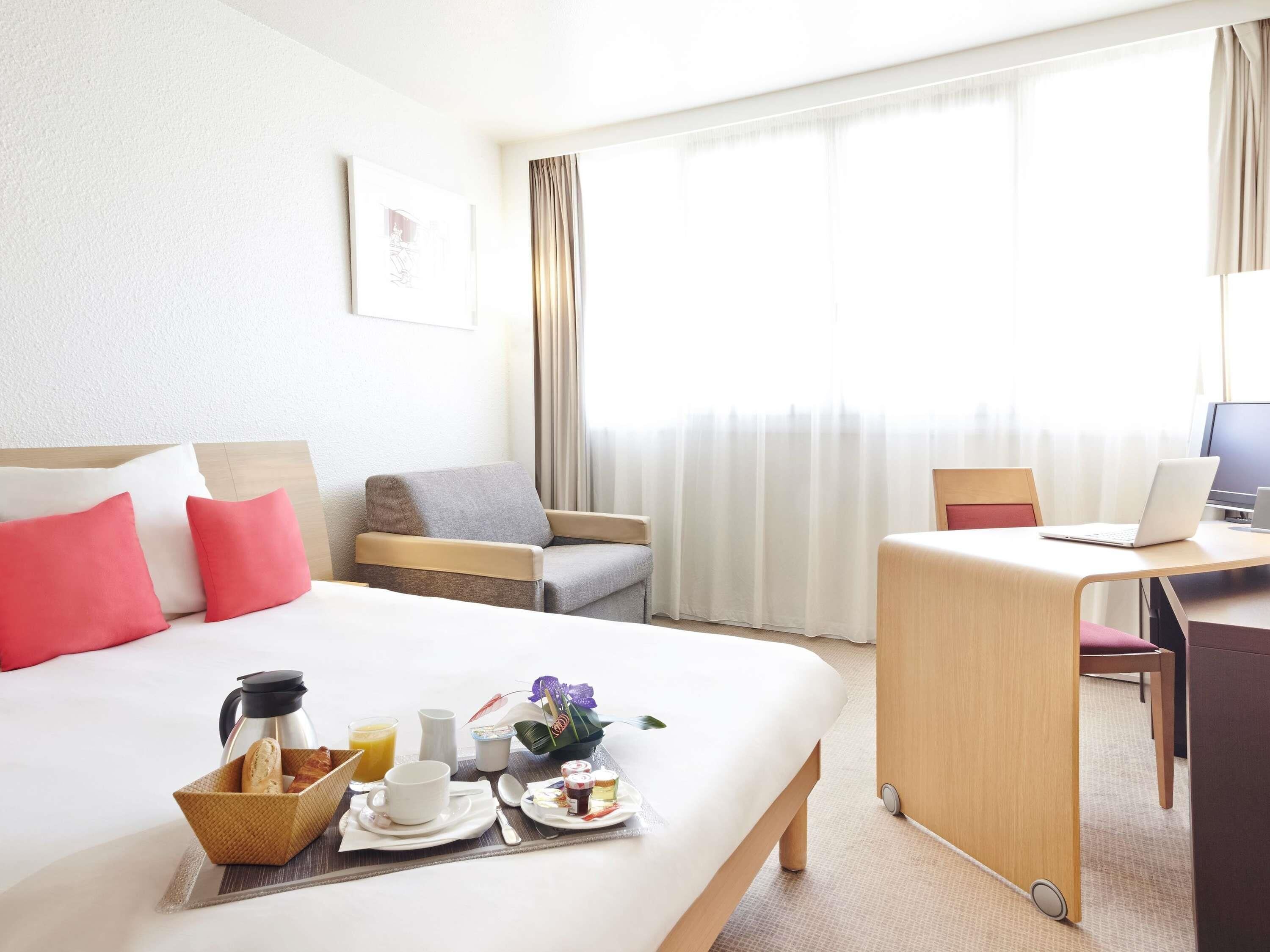 A hotel room with a bed, a desk with a laptop, a chair, and a breakfast tray with coffee, juice, and pastries.