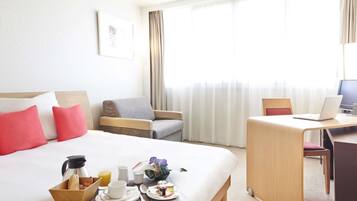 A hotel room with a bed, a desk with a laptop, a chair, and a breakfast tray with coffee, juice, and pastries.