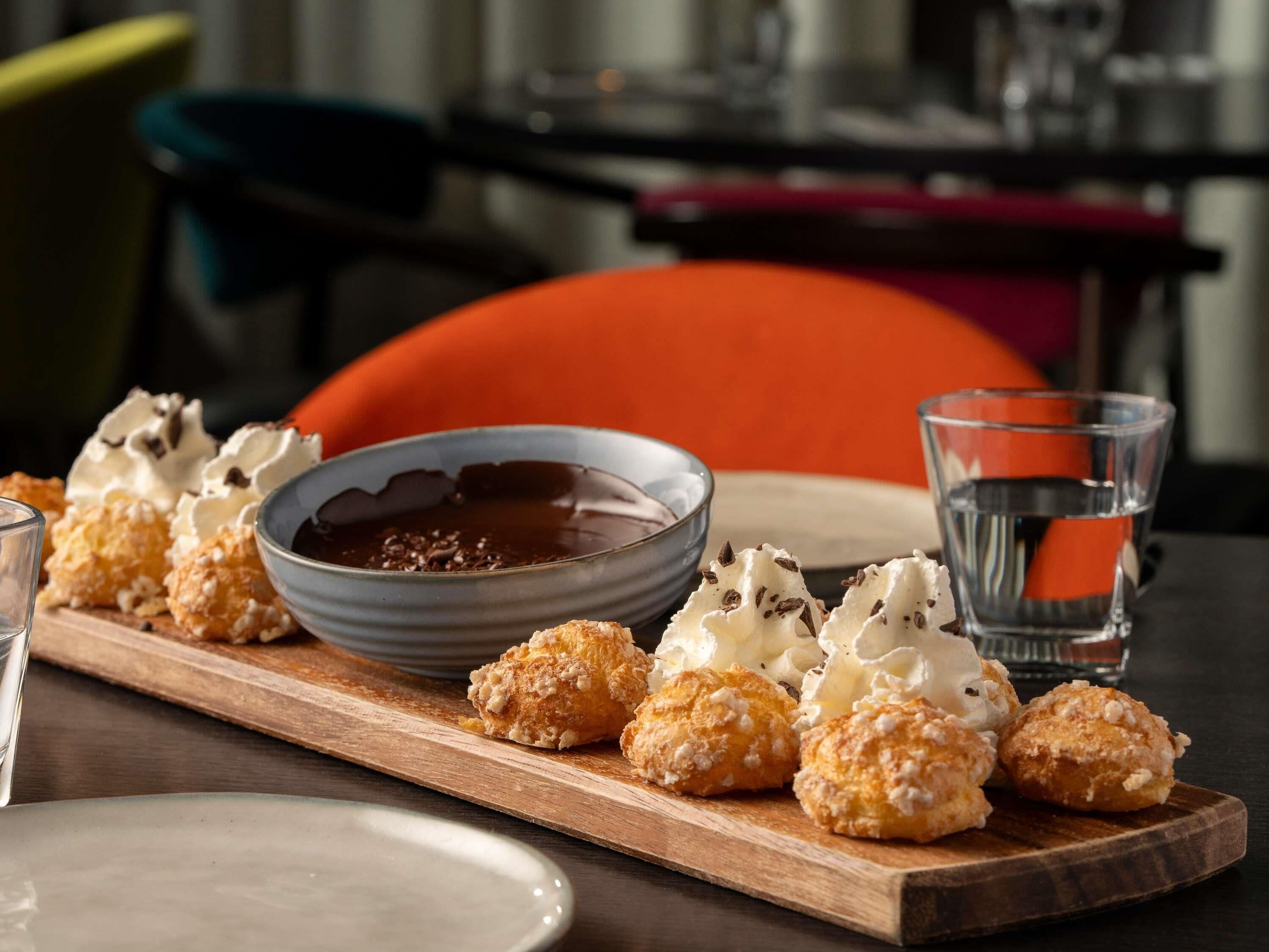 Una tabla de madera con pastelería y un bol de salsa de chocolate, un vaso de agua y un plato sobre una mesa.