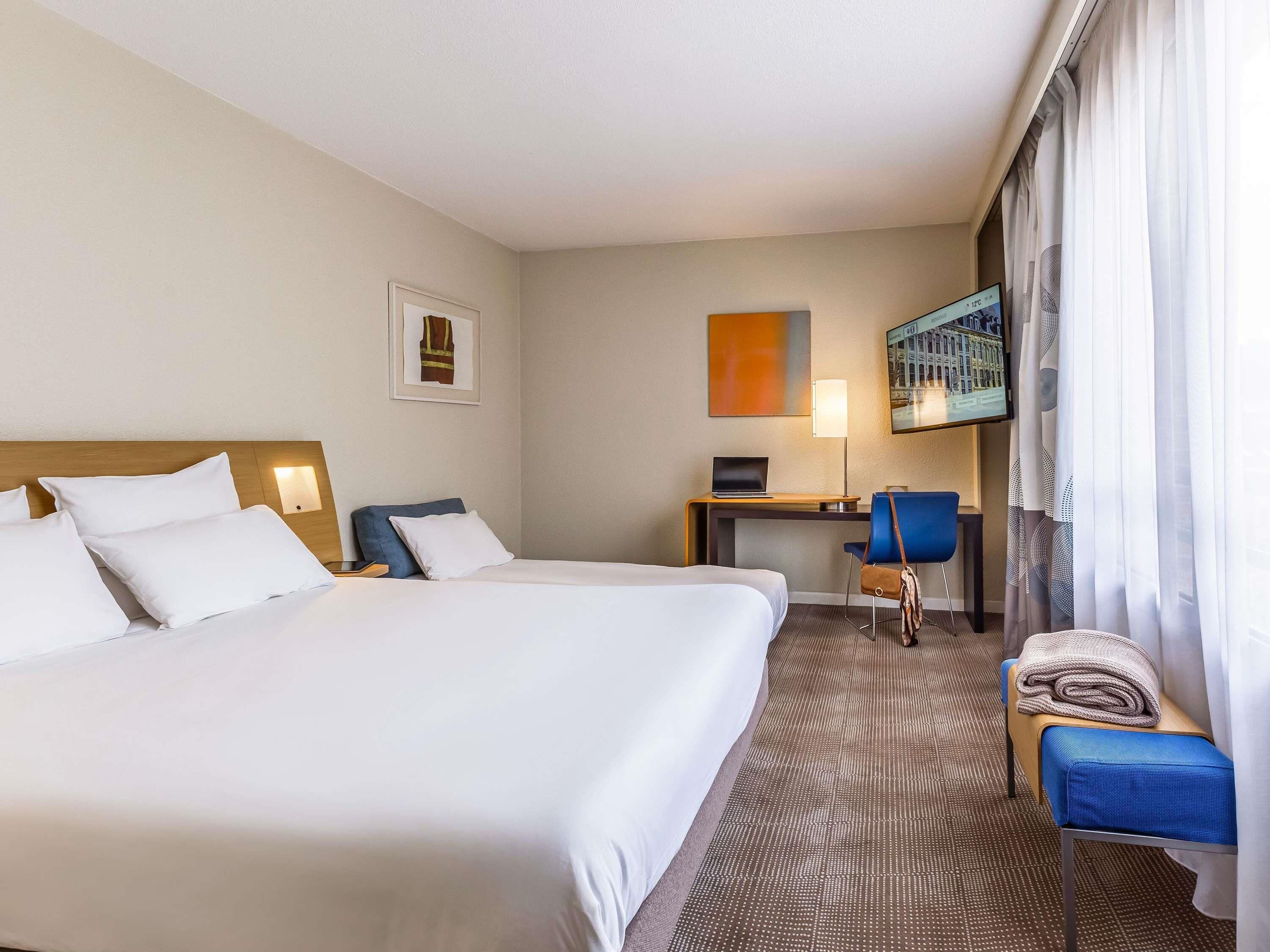 A hotel room with two beds, a desk, a chair, a TV, and a window with curtains.