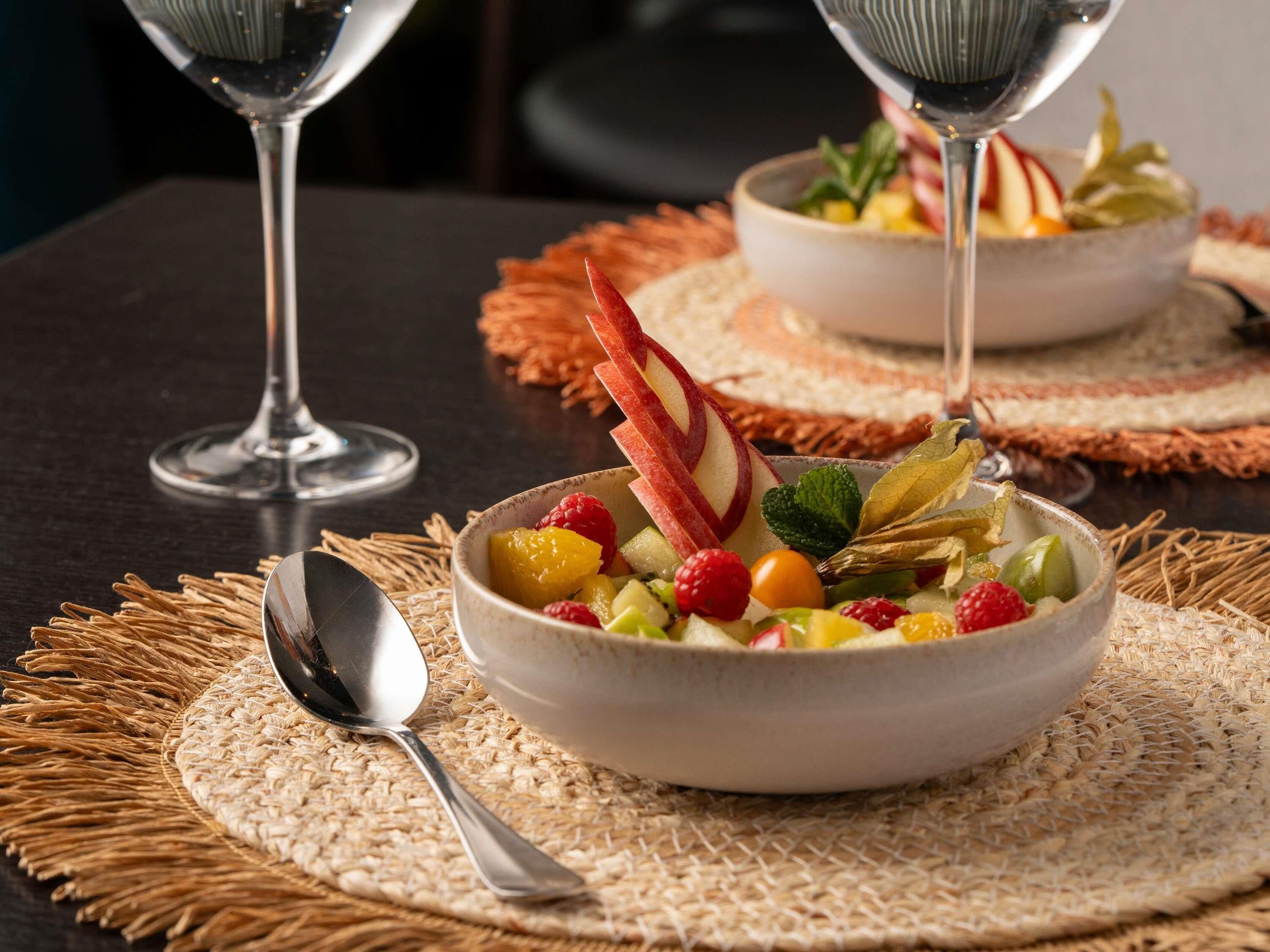 A bowl of fruit salad on a woven placemat with a spoon beside it.