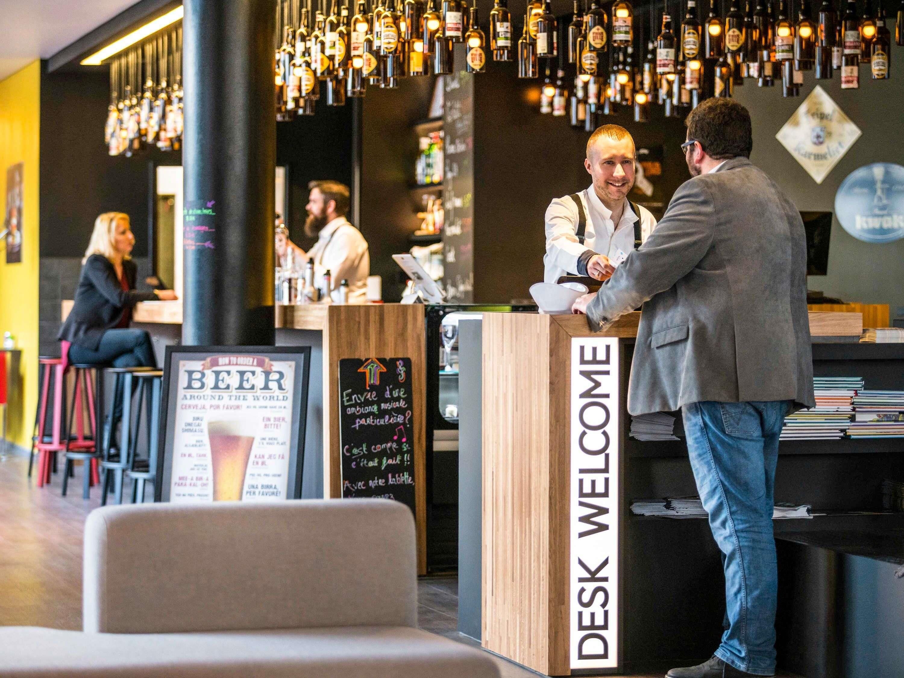 Una zona de bar con un mostrador de madera con la etiqueta 'DESK WELCOME', una pizarra con escritura y un cartel de 'BEER'.