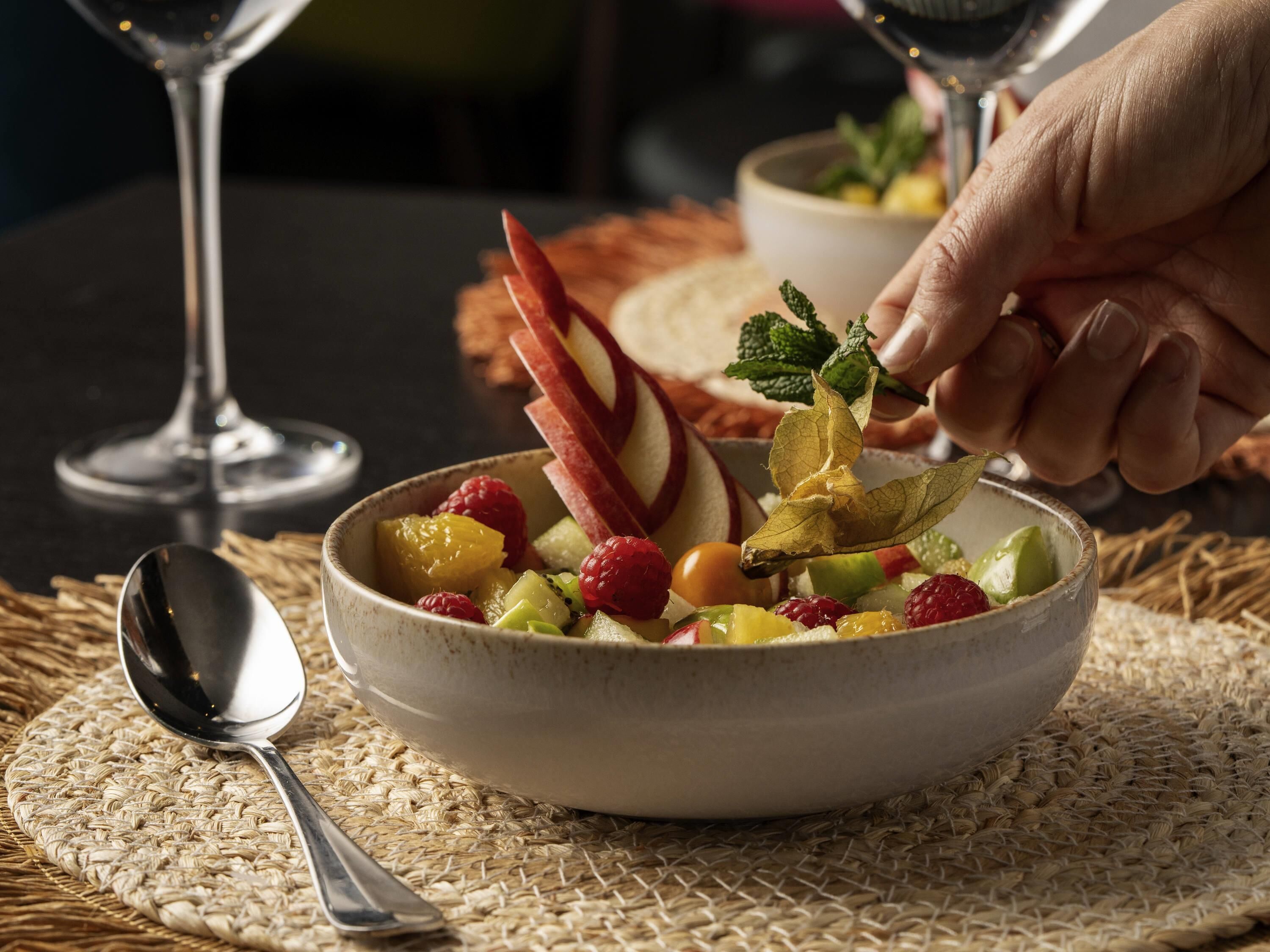 A bowl of fruit salad with a spoon and a glass of water on a woven placemat.