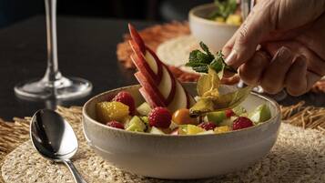 A bowl of fruit salad with a spoon and a glass of water on a woven placemat.