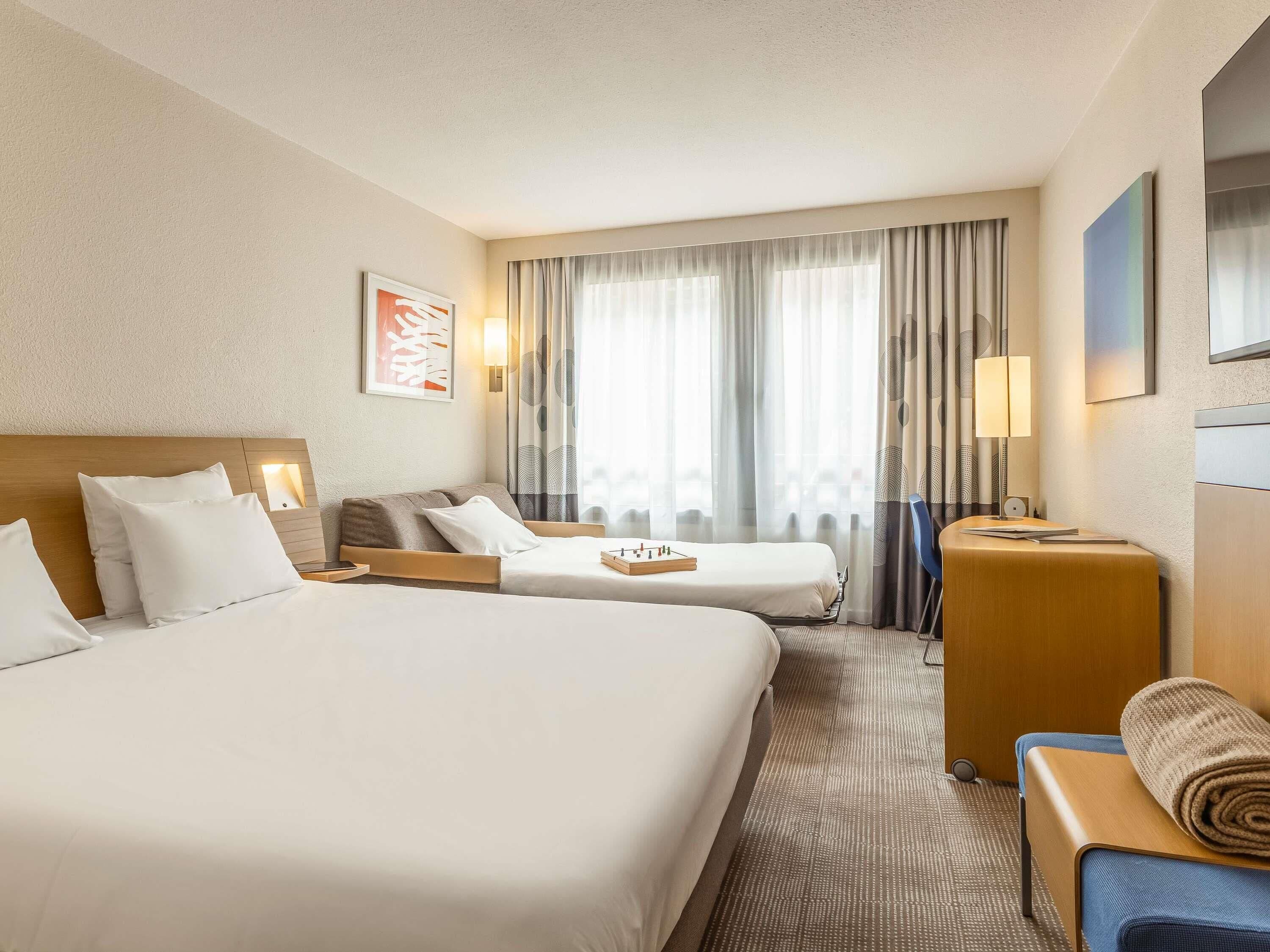 Hotelkamer met twee bedden, een bureau, een stoel, een lamp, een televisie en een raam met gordijnen.