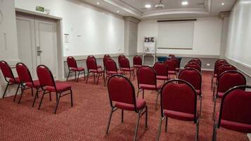 Un salón de conferencias con filas de sillas rojas, suelo alfombrado, paredes blancas y una pantalla para proyector.