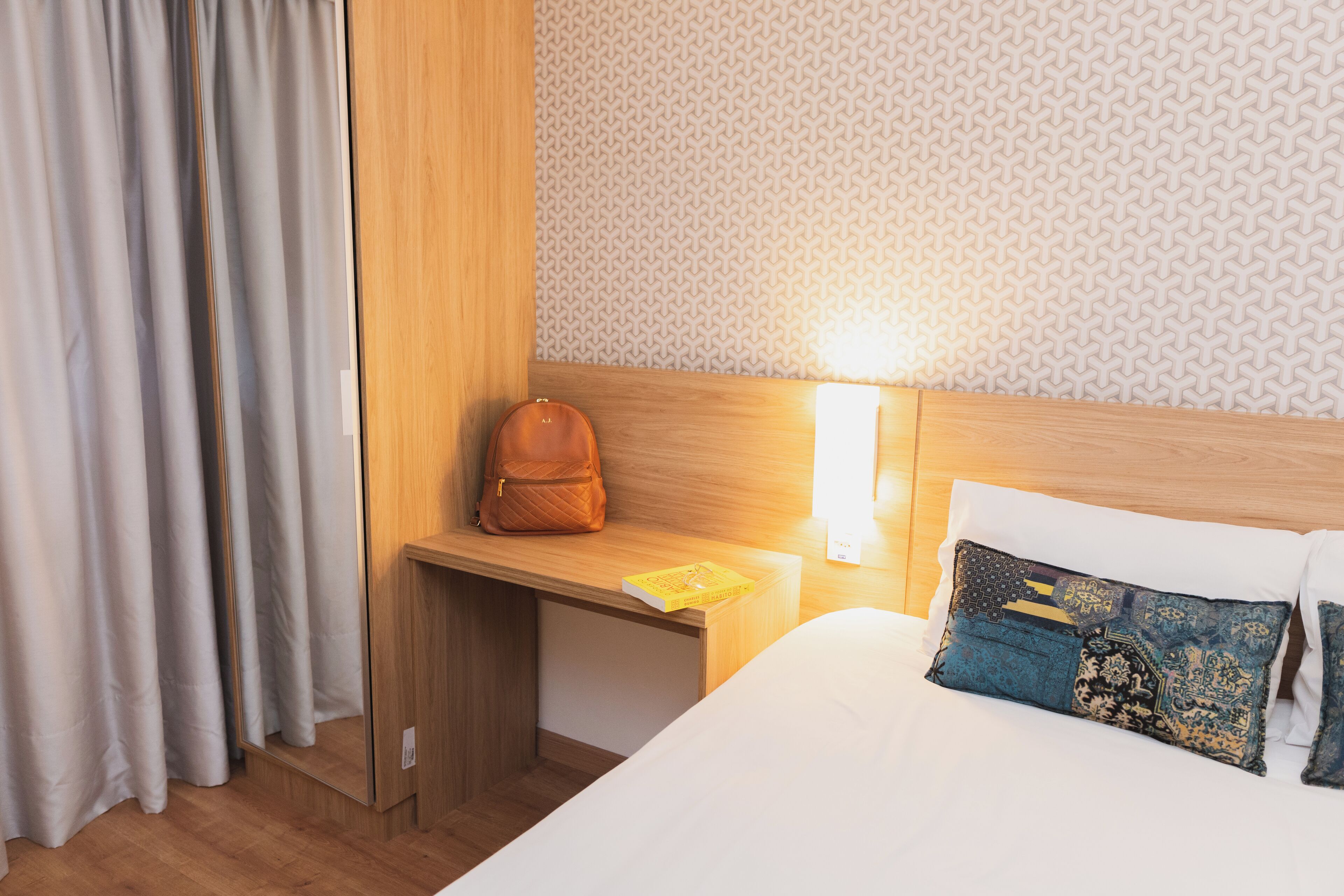 A hotel room with a wooden headboard, a bedside table with a lamp, a bed with decorative pillows, and a window with curtains.