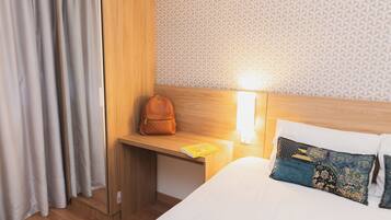 A hotel room with a wooden headboard, a bedside table with a lamp, a bed with decorative pillows, and a window with curtains.
