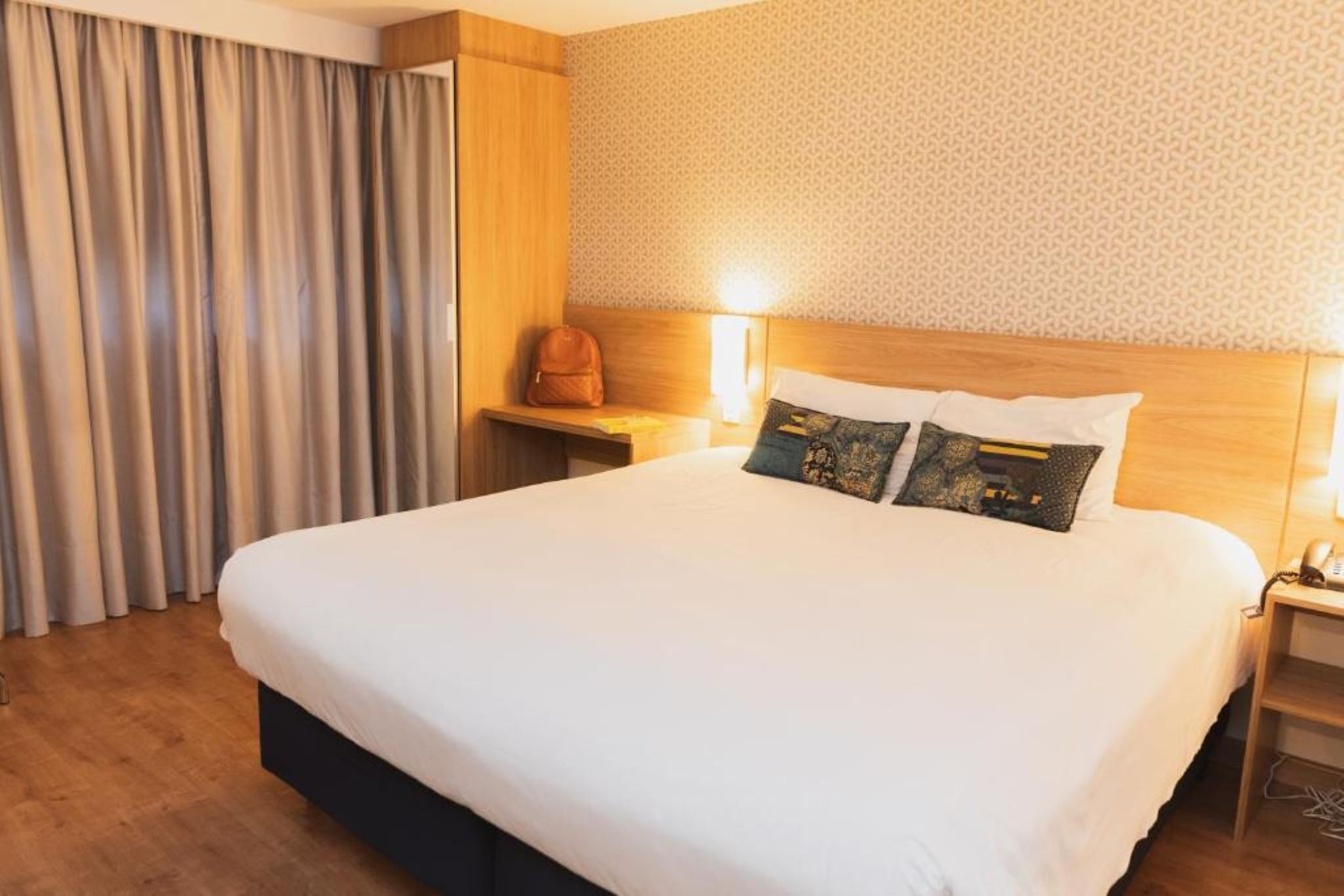 A hotel room with a large bed, a wooden headboard, a nightstand with a lamp, a suitcase, and a patterned wall.