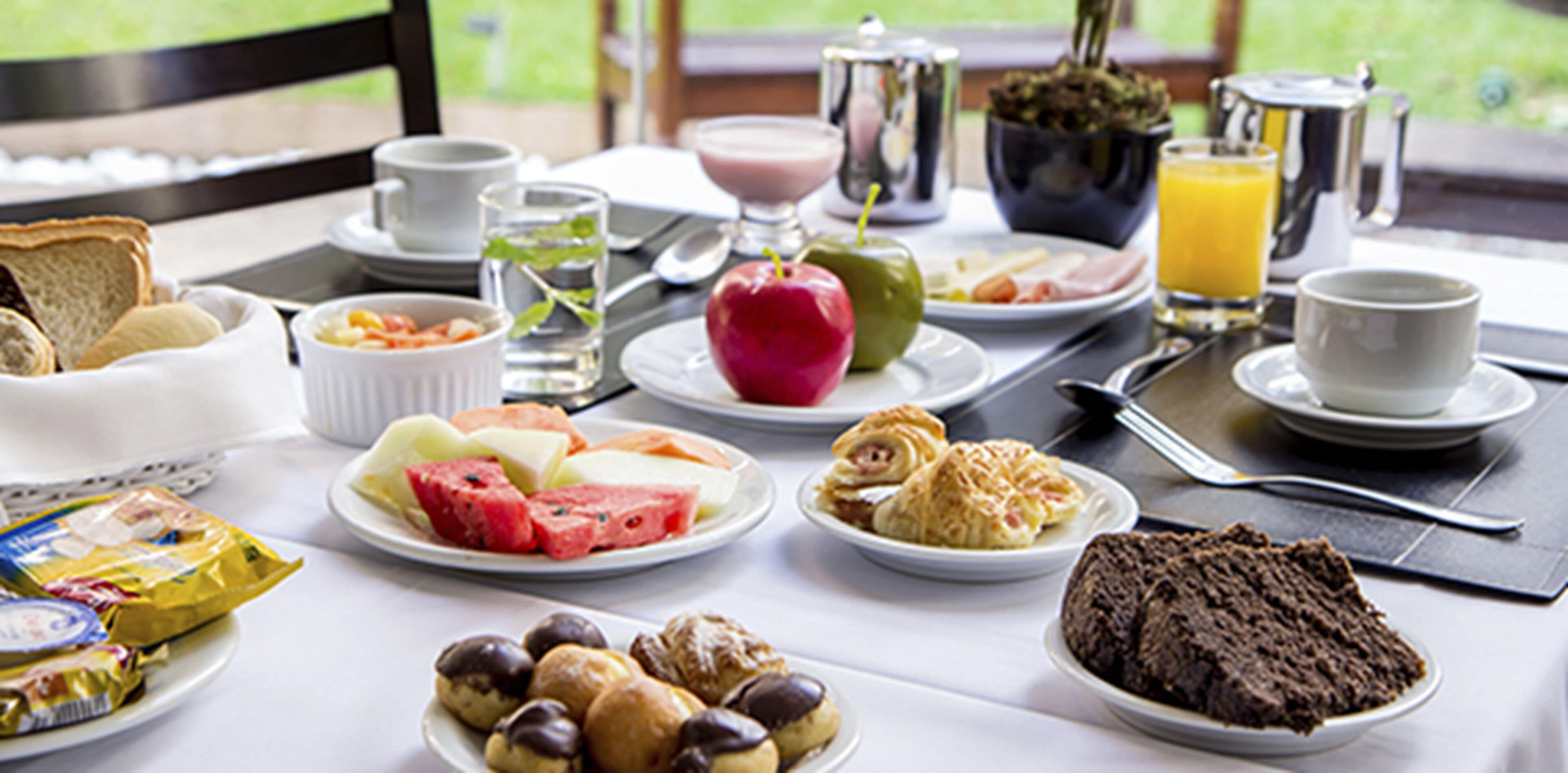 Una mesa de desayuno con diversos alimentos, como pan, frutas, bollería y una bebida caliente.