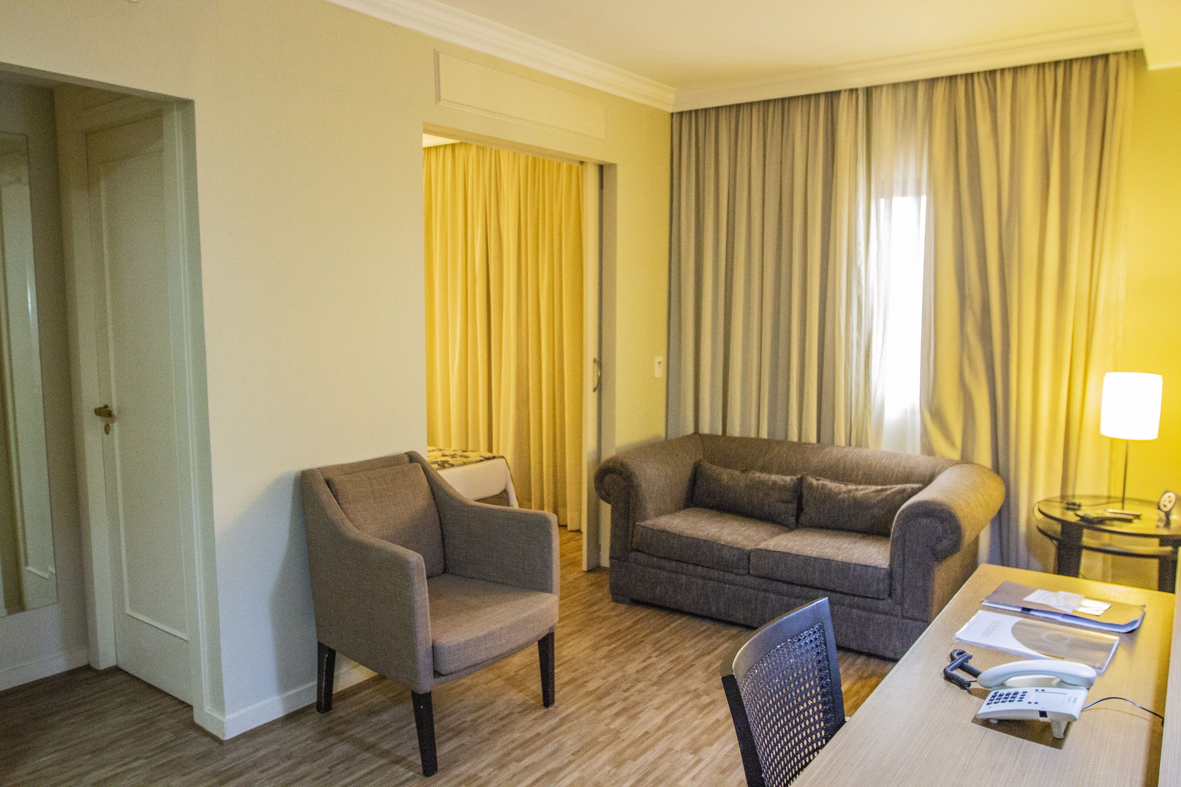 Una habitación de hotel con piso de madera, un sillón gris, un sofá gris, una mesa de comedor con una silla y una lámpara sobre una mesita de noche.