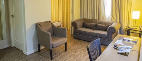 A hotel room with a wooden floor, a grey armchair, a grey sofa, a dining table with a chair, and a lamp on a side table.
