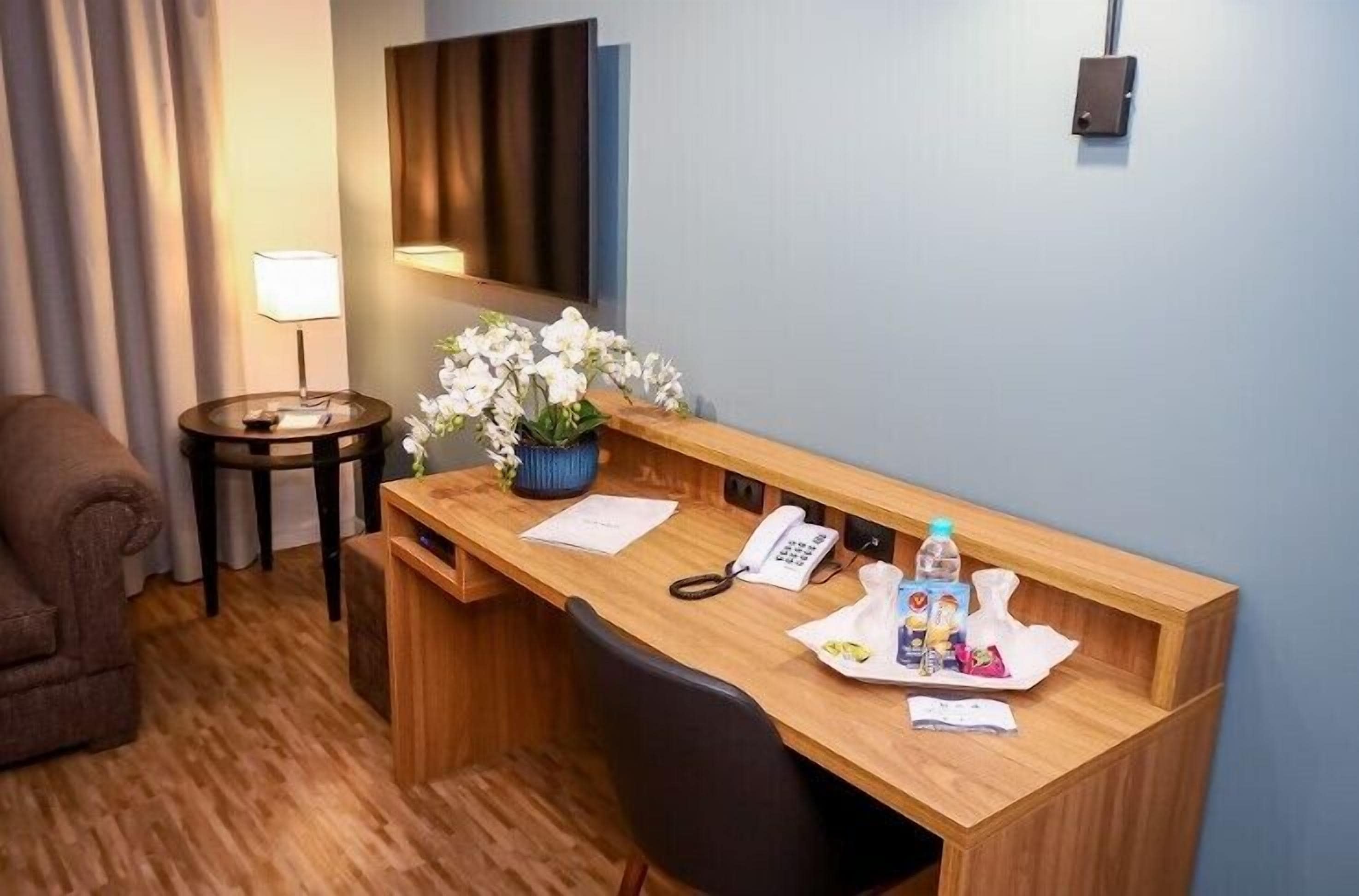 Een hotelkamer met een houten bureau, een telefoon, een lamp, een kleine plant en een waterfles.
