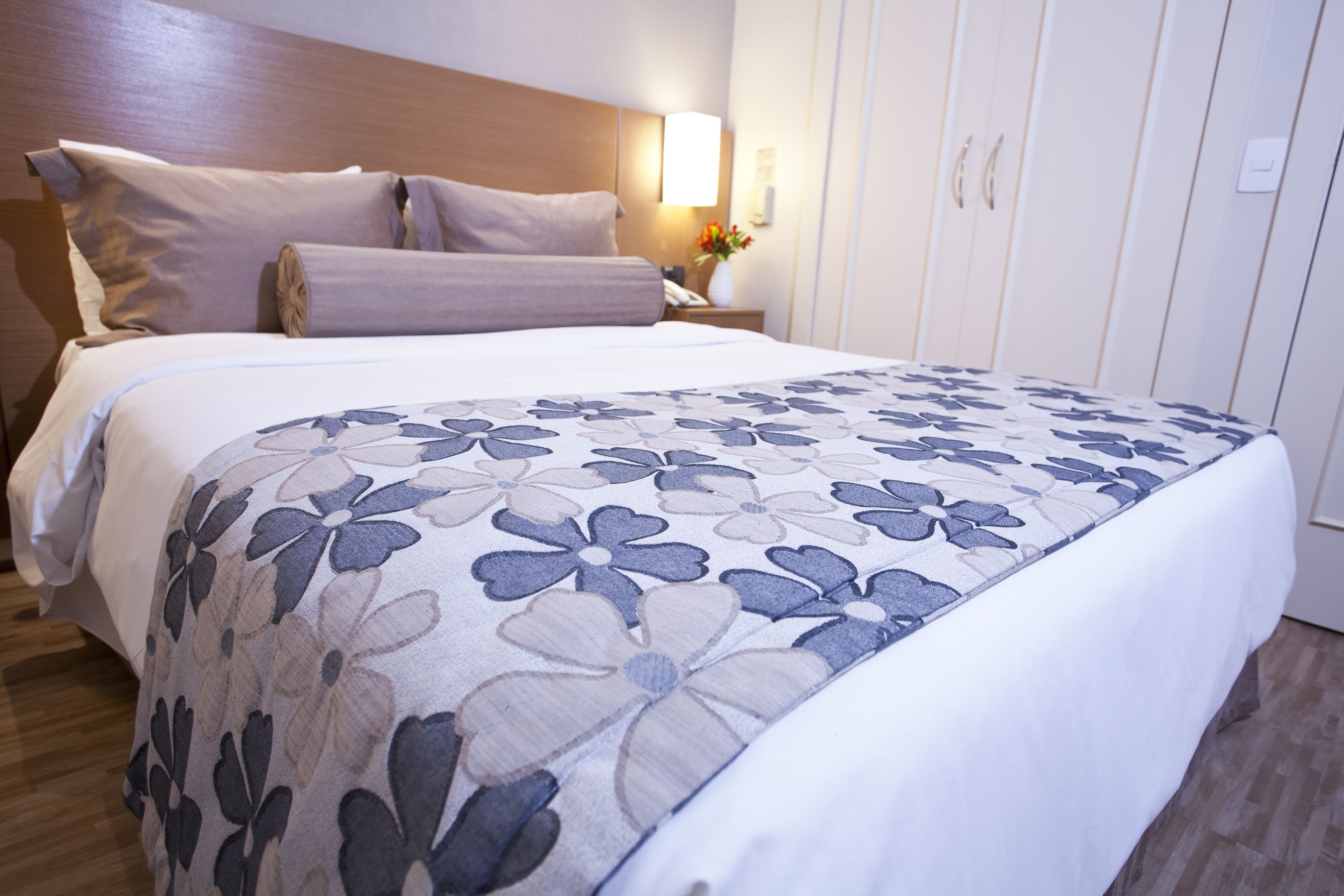 A neatly made bed with a floral-patterned bedspread, a wooden headboard, and a bedside table with a lamp and vase of flowers.