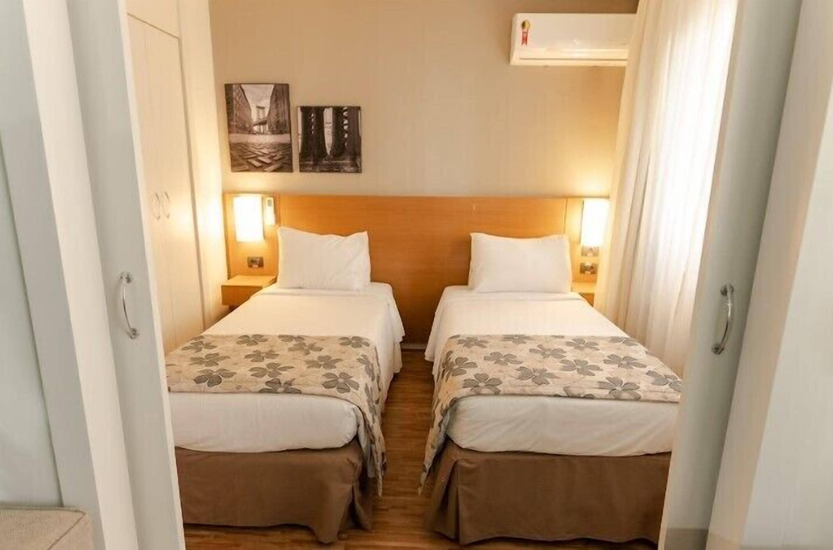 A hotel room with two beds, each with a wooden headboard, white bedding, and a floral-patterned duvet. There are wall-mounted lamps on either side of the beds, and a small table with a lamp between them.