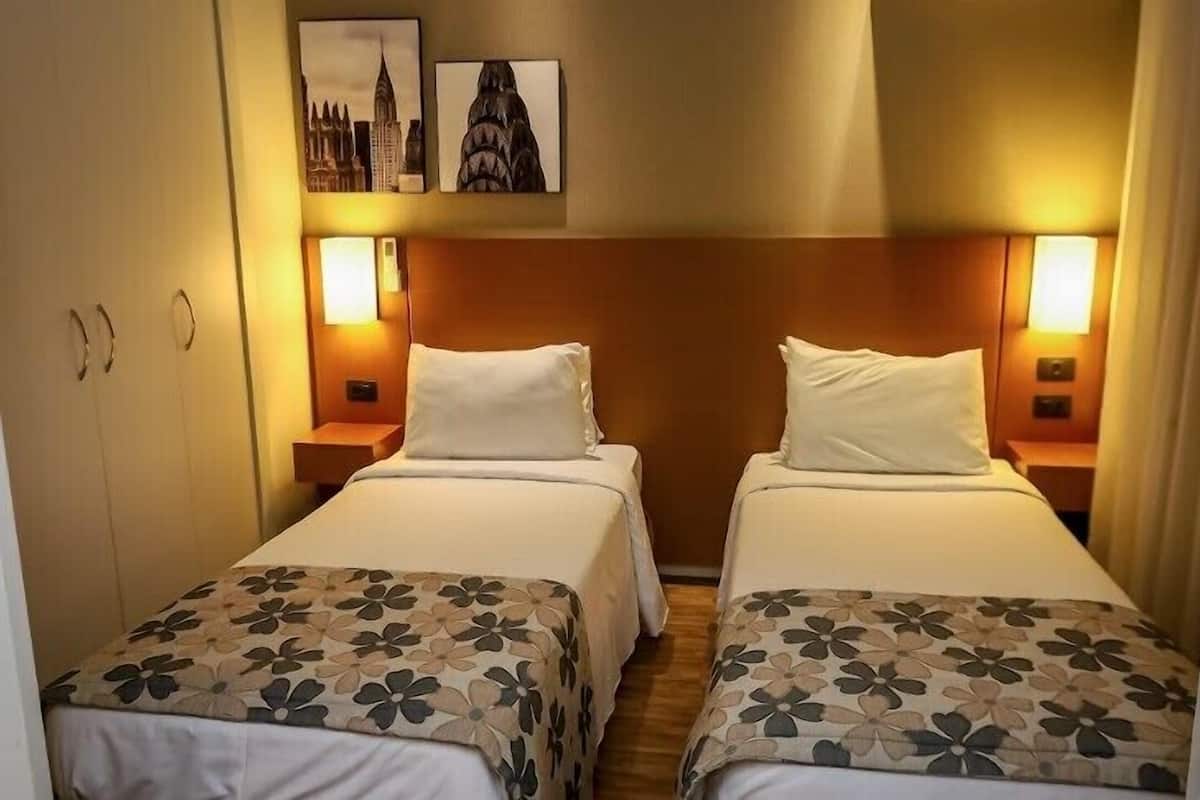 A hotel room with two single beds, each with a floral-patterned bedspread and white pillows. There are two bedside tables with lamps, and the walls are adorned with framed pictures.