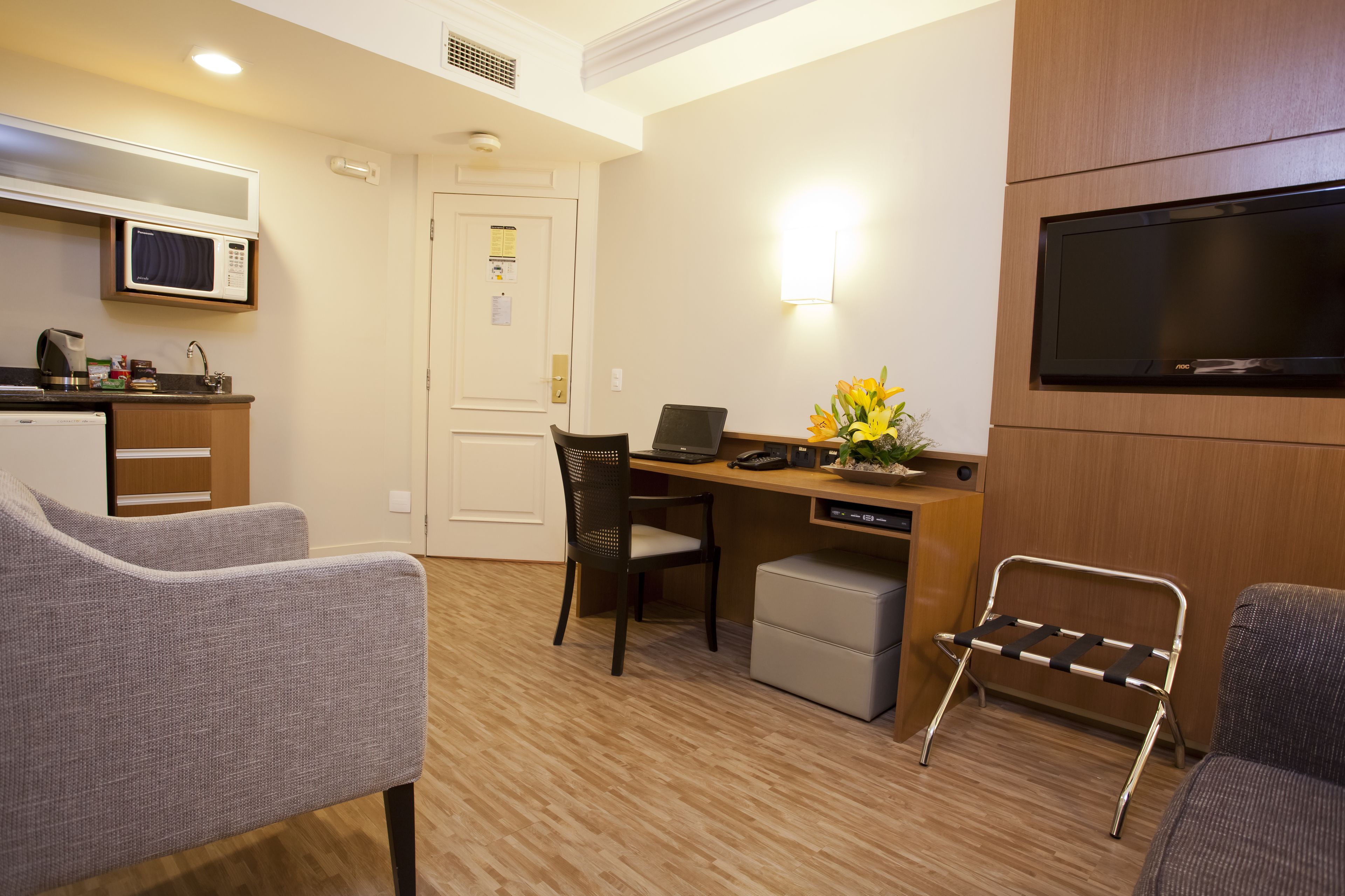A compact hotel room with a kitchenette, a desk with a laptop, a TV, and two chairs.