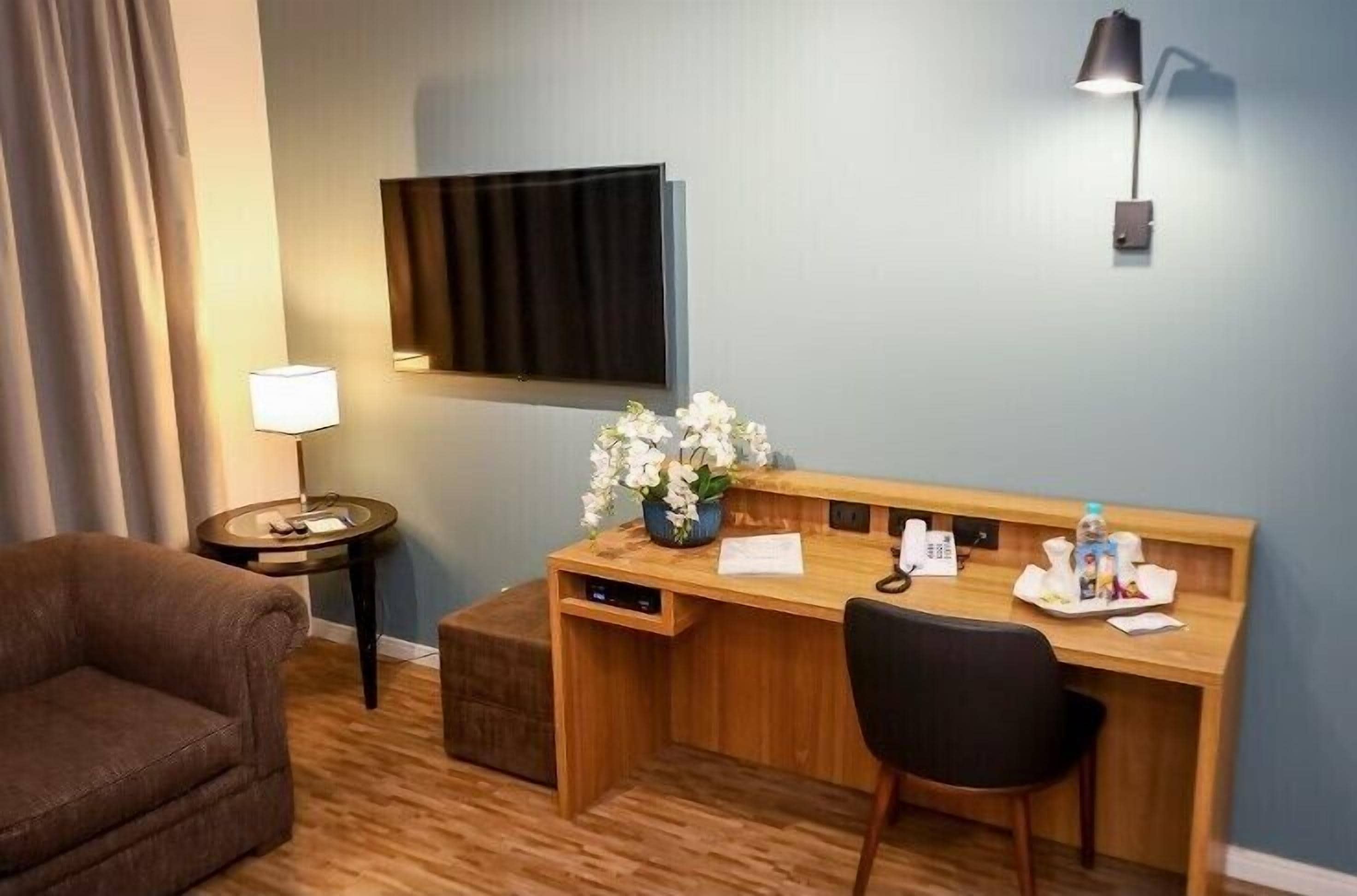 Ein Hotelzimmer mit Holzschreibtisch, Wandfernseher, Lampe, Blumenvase und braunem Sofa.