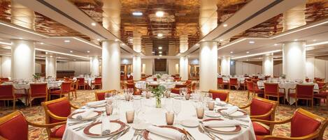 Un salón de banquetes con mesas redondas dispuestas para comer, sillas tapizadas en rojo y un techo decorativo.