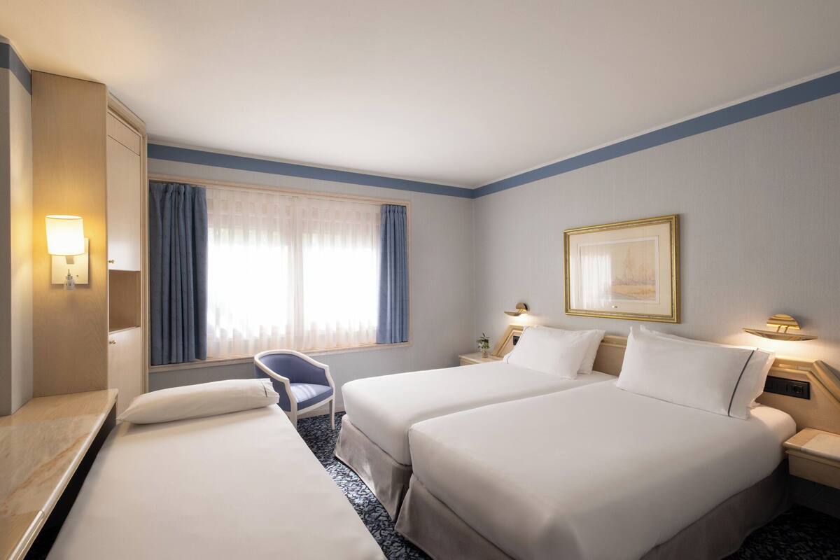 A hotel room with two beds, a desk, a chair, a window with curtains, and a framed picture on the wall.