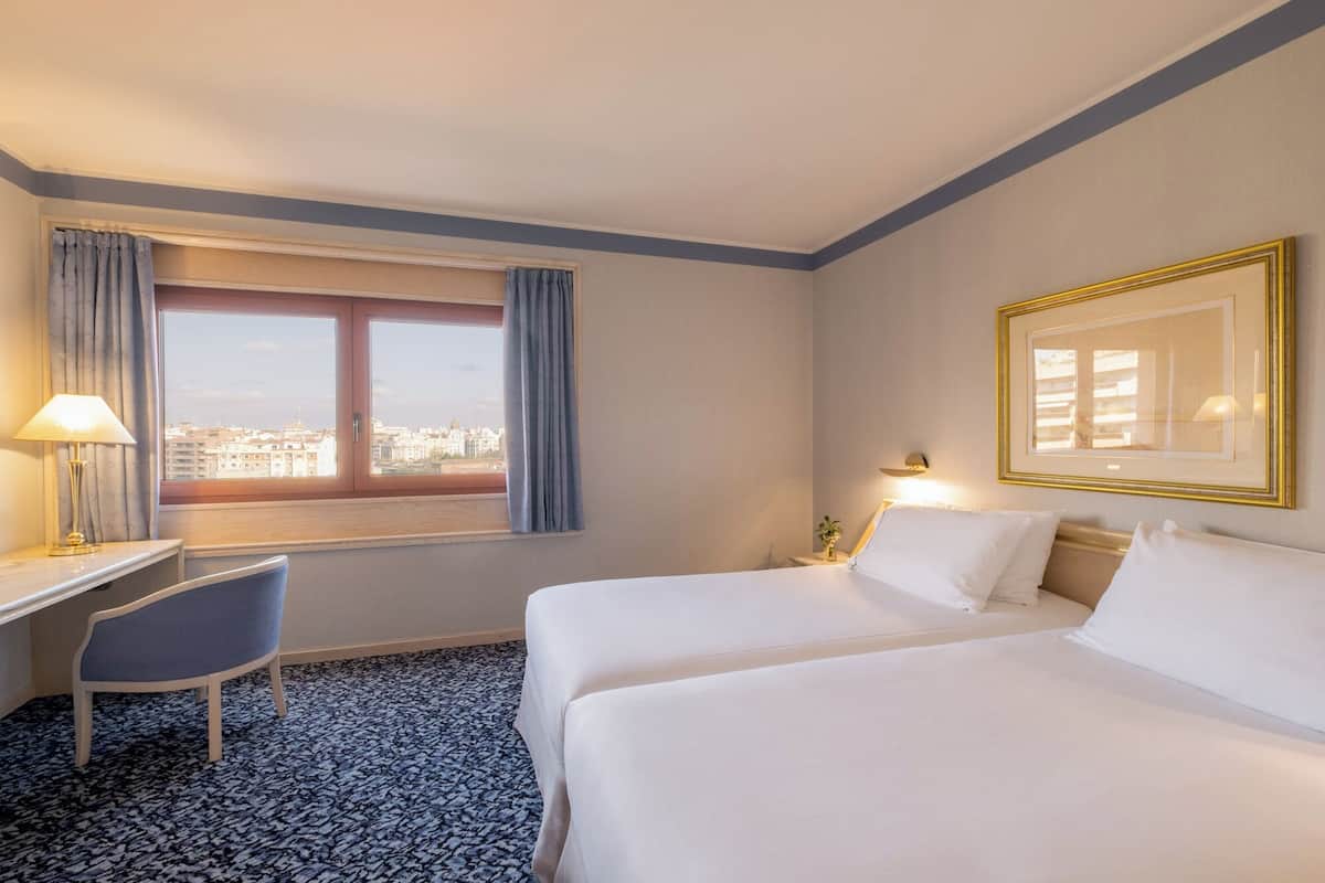 A hotel room with two beds, a desk, a chair, a window with curtains, and a framed picture on the wall.