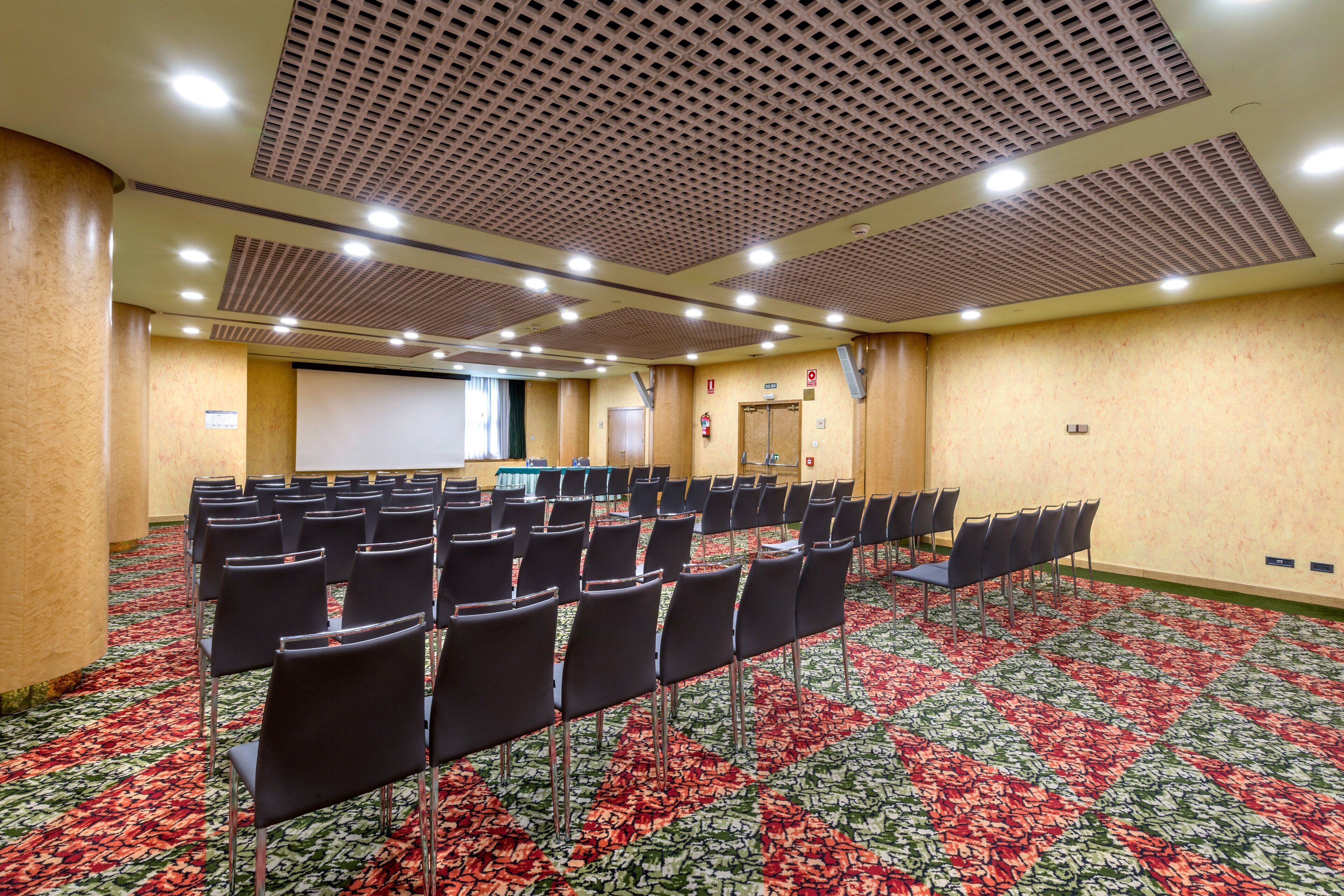 Una sala conferenze con file di sedie, uno schermo per proiettore e un pavimento rivestito di moquette.