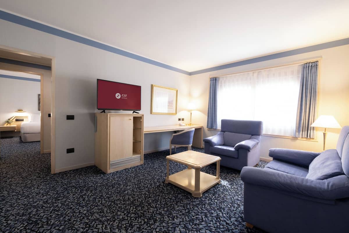 A hotel room with a TV, a desk, two chairs, a sofa, and a window with curtains.