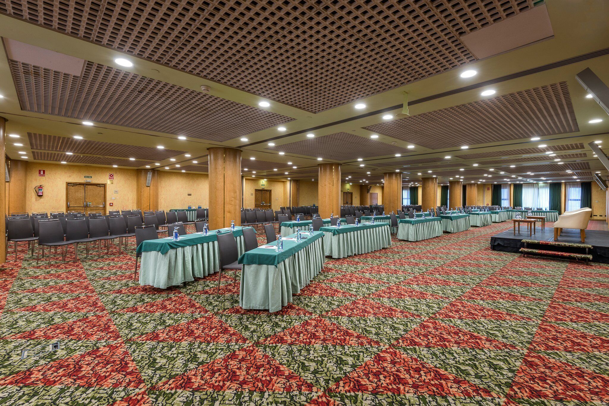 A large conference room with rows of tables and chairs, a patterned carpet, and a high ceiling with ventilation.