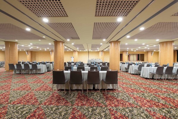 Un amplio salón de conferencias con varias mesas y sillas, piso alfombrado y conductos de ventilación en el techo.