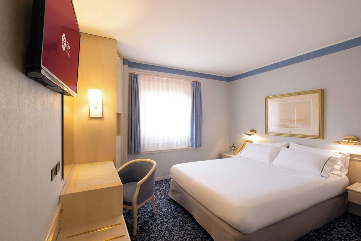 A hotel room with a bed, bedside tables, a desk, a chair, a TV, and a window with curtains.