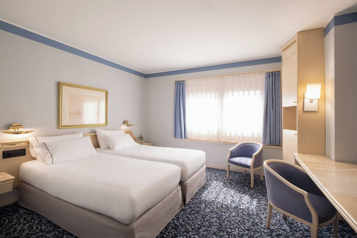 A hotel room with two beds, a desk, a chair, a window with curtains, and a framed picture on the wall.